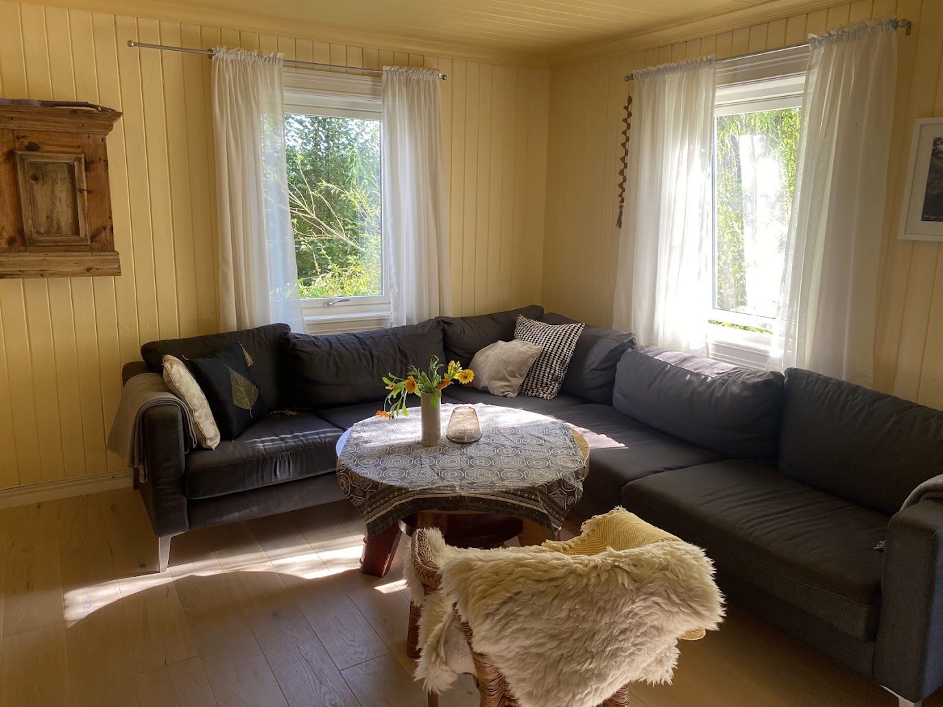 Stuen i gjestehuset med sofa, skinn og vedovn, og lyse gardiner i vinduene